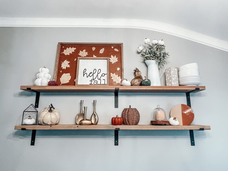May include: Two wooden shelves with black metal brackets, decorated with fall-themed items, including pumpkins, gourds, a white pitcher, and a sign that reads "hello fall".