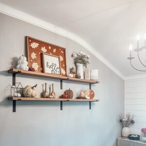 May include: Two wooden shelves with black metal brackets, decorated with fall-themed items, including pumpkins, gourds, leaves, and a sign that says "hello fall".