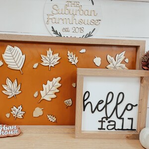 May include: A wooden sign with the text "The Suburban Farmhouse" est. 2018. A brown frame with a tan background and wooden cutouts of leaves and acorns. A white sign with the text "hello fall" in black cursive lettering.