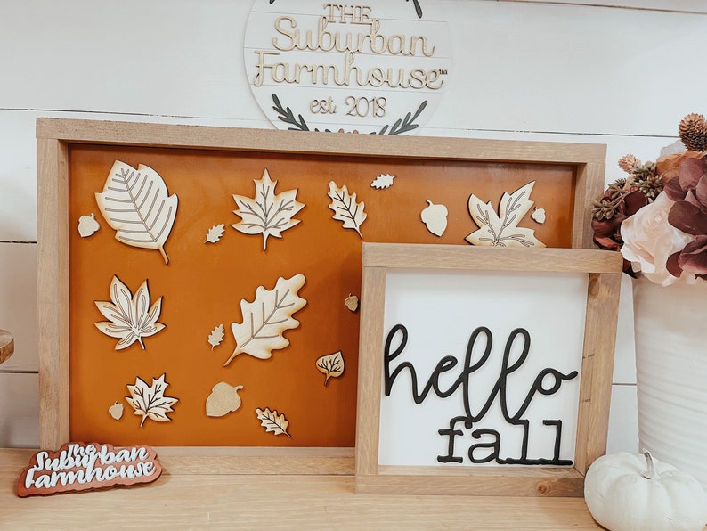 May include: A wooden sign with the text "The Suburban Farmhouse" est. 2018. A brown frame with a tan background and wooden cutouts of leaves and acorns. A white sign with the text "hello fall" in black cursive lettering.