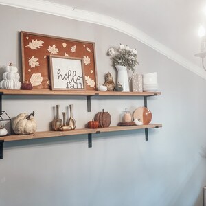 May include: Two wooden shelves with black metal brackets, decorated with fall-themed items, including a sign that says "hello fall", pumpkins, gourds, and a white ceramic vase with cotton stems.