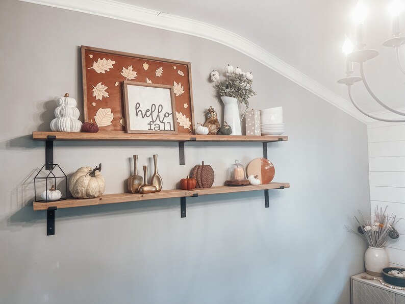 May include: Two wooden shelves with black metal brackets, decorated with fall-themed items, including a sign that says "hello fall", pumpkins, gourds, and a white ceramic vase with cotton stems.