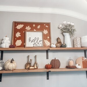 May include: Two wooden shelves with black metal brackets, decorated with fall-themed items. The top shelf has a framed sign that says "hello fall", a framed picture with fall leaves, a white ceramic pumpkin, a small brown and gold gourd, a white ceramic pumpkin, and a green gourd. The bottom shelf has a black metal lantern with a white ceramic pumpkin inside, a white ceramic pumpkin, a gold metal vase, a gold metal vase, a gold metal vase, a small orange pumpkin, a brown and gold pumpkin, a glass cloche with a white ceramic pumpkin inside, and a brown and orange plate.