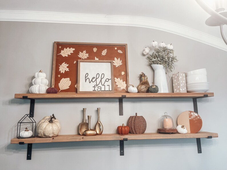 May include: Two wooden shelves with black metal brackets, decorated with fall-themed items. The top shelf has a framed sign that says "hello fall", a framed picture with fall leaves, a white ceramic pumpkin, a small brown and gold gourd, a white ceramic pumpkin, and a green gourd. The bottom shelf has a black metal lantern with a white ceramic pumpkin inside, a white ceramic pumpkin, a gold metal vase, a gold metal vase, a gold metal vase, a small orange pumpkin, a brown and gold pumpkin, a glass cloche with a white ceramic pumpkin inside, and a brown and orange plate.