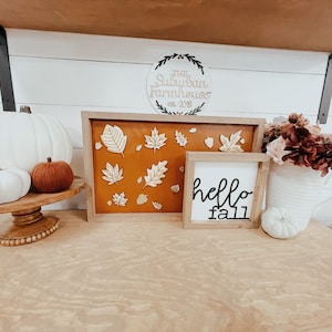 May include: A wooden shelf with fall decor, including white pumpkins, a brown and white pumpkin, a wooden sign that says "The Suburban Farmhouse est. 2018", a framed piece of art with gold leaves on a brown background, and a white sign that says "hello fall".