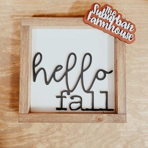 May include: A wooden sign with a white background and black lettering that says "hello fall". The sign is framed in a natural wood frame. A small wooden sign with the text "The Suburban Farmhouse" is in the top right corner of the image.