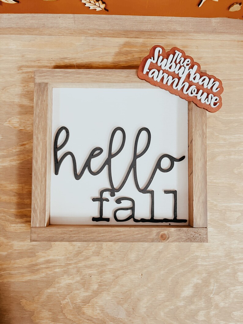 May include: A wooden sign with a white background and black lettering that says "hello fall". The sign is framed in a natural wood frame. A small wooden sign with the text "The Suburban Farmhouse" is in the top right corner of the image.