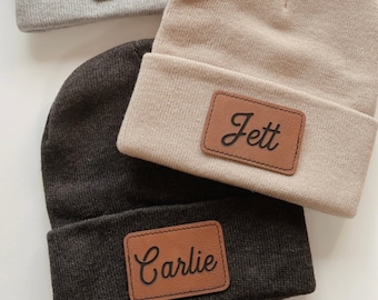 May include: Three knit beanies with personalized leather patches. The beanies are gray, beige, and black. The patches have the names "Christina", "Jett", and "Carlie" written in cursive.