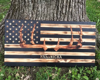 May include: A wooden American flag with a black background and a burnt wood effect. The flag has a pair of antlers in the center and the name "Kvasnicka" written below the antlers.
