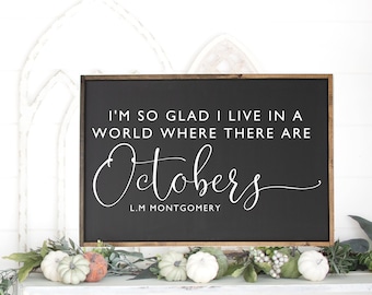 May include: Black wooden sign with white text that reads "I'm so glad I live in a world where there are Octobers" with the author L.M. Montgomery below. The sign is sitting on a white shelf with a garland of green leaves and white pumpkins.