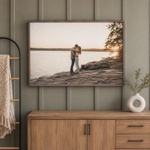 May include: A rectangular framed wall art print featuring a photograph of two people embracing on a rocky shoreline by a lake at sunset. The image displays a calm body of water, distant hills, and a tree silhouette under a warm, golden sky.