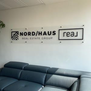 May include: A clear acrylic sign with black lettering that reads "Nord/Haus Real Estate Group". The sign is mounted on a white wall above a blue leather sectional sofa.