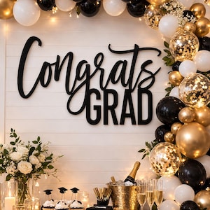 May include: A graduation party scene with a black table, balloons, and decorations. The words "Congrats GRAD" are displayed in large black letters. Gold, black, and white balloons create an arch. Champagne, cupcakes, and candles complete the festive setting.