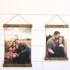 May include: Two framed photo prints hanging on a white wall. Each print is held by a wooden frame and suspended by twine. The photos depict a couple in a field and a couple embracing. The wooden frames have a rustic, natural finish.