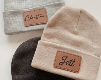 May include: Three knit beanies in gray, tan, and dark brown. Each beanie has a rectangular brown leather patch with a name in cursive: "Christina", "Jett", and "Carlie". The beanies are folded at the brim.