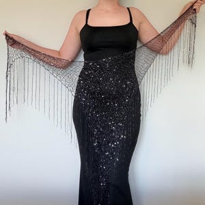 Może przedstawiać: Czarna suknia wieczorowa z błyszczącym, cekinowym stanikiem i pasującą, przezroczystą szalem z koralikowymi frędzlami. Sukienka ma dopasowaną sylwetkę i ramiączka spaghetti. Szal jest trzymany na boki.