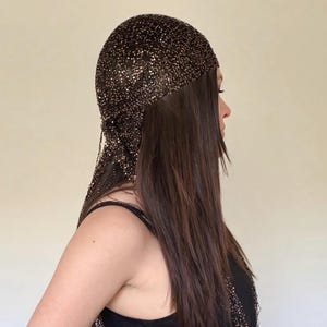 Pode incluir: Um adorno de cabeça marrom escuro com contas com um design drapeado. O adorno de cabeça é feito de pequenas contas reflexivas e é usado sobre a cabeça e o cabelo. A pessoa está usando um top preto.