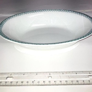 May include: White oval serving bowl with a teal patterned border. The bowl has a slightly raised base and a smooth, glossy surface. A ruler is in the foreground, providing a sense of scale. This bowl is suitable for serving dishes.