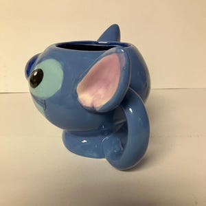 May include: Blue ceramic mug shaped like Stitch from Lilo & Stitch. The mug has a pink ear and a blue tail that forms the handle.