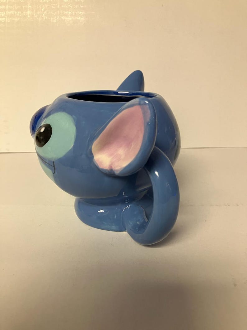 May include: Blue ceramic mug shaped like Stitch from Lilo & Stitch. The mug has a pink ear and a blue tail that forms the handle.
