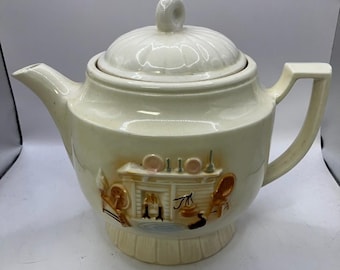 May include: Cream-colored ceramic teapot with a lid and handle. The teapot features a decorative relief design of a fireplace scene with chairs and a table. The spout and handle are smooth, while the lid has a ridged texture.