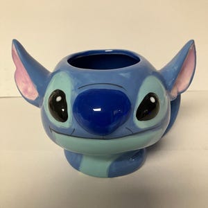 May include: Blue ceramic mug shaped like Stitch from Lilo & Stitch. The mug has a light blue, dark blue, and pink color scheme. Stitch has a big smile and black eyes.