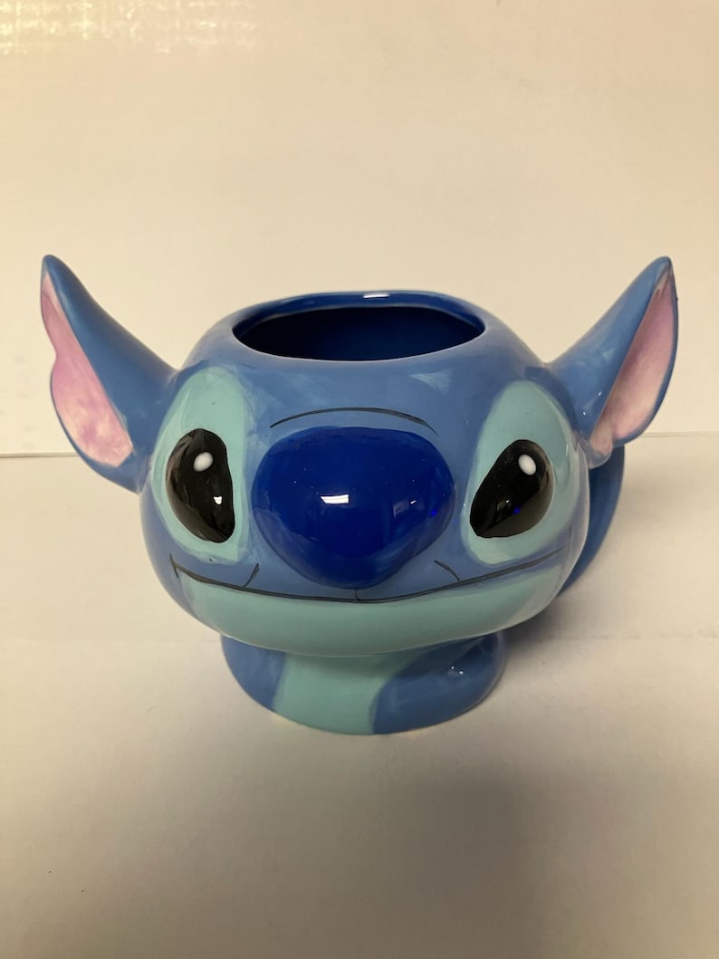 May include: Blue ceramic mug shaped like Stitch from Lilo & Stitch. The mug has a light blue, dark blue, and pink color scheme. Stitch has a big smile and black eyes.