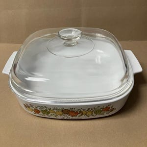 May include: White casserole dish with a clear glass lid. The dish has a square shape with rounded corners and white handles. A colorful border of fruits and vegetables decorates the dish. The text "Le Romarin" is printed on the dish.