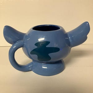 May include: Blue ceramic mug shaped like Stitch from Lilo & Stitch. The mug has a handle and a blue and green design.