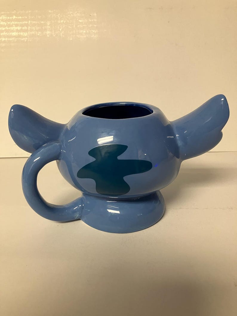 May include: Blue ceramic mug shaped like Stitch from Lilo & Stitch. The mug has a handle and a blue and green design.