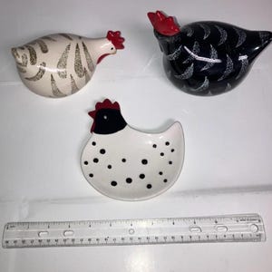 May include: Three ceramic chicken figurines. One is white with brown markings, another is black with white markings, and the third is a white dish with black spots. Each has a red comb. A ruler is at the bottom.