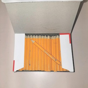 May include: A box of yellow pencils with silver metal bands and pink erasers. One pencil is angled across the others, displaying the text "UNIVERSAL 24264". The box is open, revealing the pencils inside.