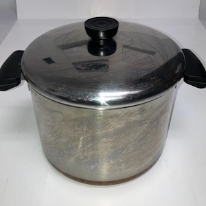 May include: A stainless steel cooking pot with a lid and two black handles. The lid has a black knob. The pot is cylindrical and appears to be used. The pot is designed for cooking.