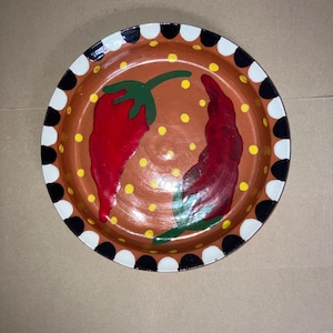 May include: Hand-painted ceramic plate with a brown interior featuring two red chili peppers and yellow polka dots. The rim has a pattern of alternating black and white semicircles. A decorative piece for kitchen or dining.