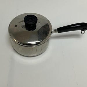 May include: A small, stainless steel saucepan with a lid and a black handle. The lid has a black knob. The handle is attached to the side of the pan and has a loop at the end. The pan is sitting on a white surface.