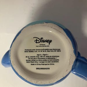 May include: Blue ceramic mug with a Disney logo and text that reads "Produced under license by/Produite sous licence par Silver Buffalo, LLC. 141 W. 36 St., New York, NY 10018 Age 8+/Age 8+ 20 oz. ceramic mug./591 ml. tasse en c&eacute;ramique. Do not use in microwave or dishwasher./Ne pas utiliser au micro-ondes ou lave-vaisselle. Hand Wash Only. / Lavage &agrave; la main seulement. Made in China./Fabriqu&eacute; en Chine. SMGL19995042018".