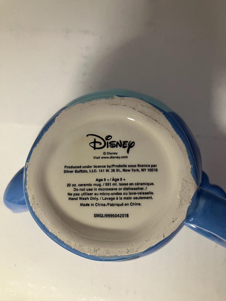 May include: Blue ceramic mug with a Disney logo and text that reads "Produced under license by/Produite sous licence par Silver Buffalo, LLC. 141 W. 36 St., New York, NY 10018 Age 8+/Age 8+ 20 oz. ceramic mug./591 ml. tasse en c&eacute;ramique. Do not use in microwave or dishwasher./Ne pas utiliser au micro-ondes ou lave-vaisselle. Hand Wash Only. / Lavage &agrave; la main seulement. Made in China./Fabriqu&eacute; en Chine. SMGL19995042018".