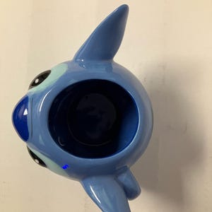 May include: Blue ceramic mug shaped like Stitch from Lilo & Stitch. The mug has a large opening and a handle on the side.