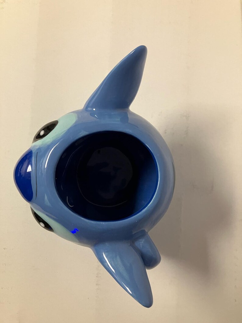 May include: Blue ceramic mug shaped like Stitch from Lilo & Stitch. The mug has a large opening and a handle on the side.