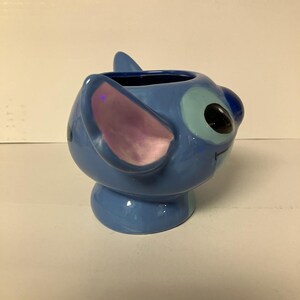 May include: A blue ceramic mug shaped like Stitch from Lilo & Stitch. The mug has a light blue glaze and a pink ear.