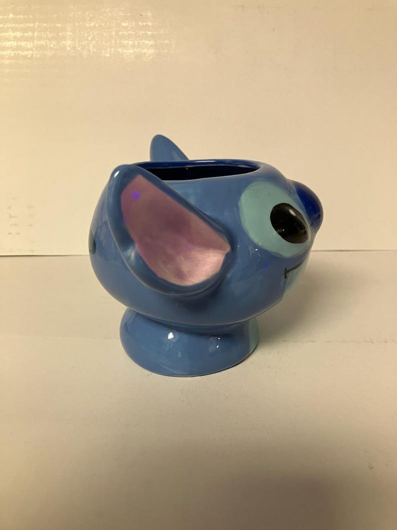 May include: A blue ceramic mug shaped like Stitch from Lilo & Stitch. The mug has a light blue glaze and a pink ear.
