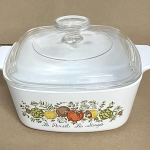 May include: White casserole dish with a clear glass lid. The dish features a colorful vegetable design with the words "Le Persil, La Sauge" in script. The dish has white handles on either side.