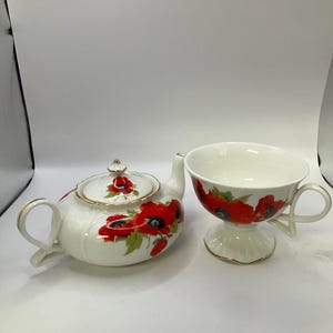 May include: A white porcelain teapot and teacup set with a red poppy floral design. The teapot has a lid and a curved handle. The teacup has a wide bowl and a handle.