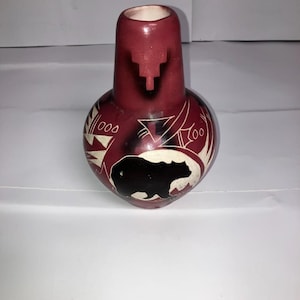 May include: A maroon-colored, handcrafted vase with a rounded body and a cylindrical neck. The vase features etched white and black designs, including a bear silhouette and geometric patterns. The vase is likely made of ceramic or a similar material.