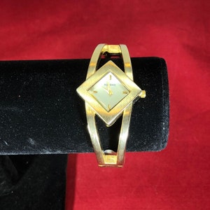 May include: A gold-coloured wristwatch featuring a diamond-shaped face and a bracelet-style band. The watch face is light-coloured with gold hands and the brand name "N.Y. Time". The watch is displayed on a black velvet stand.