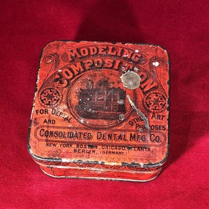 May include: A red and black square tin box with the text "MODELING COMPOSITION" in a circular design. The text "FOR DENTAL AND OTHER PURPOSES" is printed below the circular design. The text "CONSOLIDATED DENTAL MFG. CO." is printed below the text. The text "NEW YORK, BOSTON, CHICAGO,LANTA, BERLIN, GERMANY" is printed below the company name.
