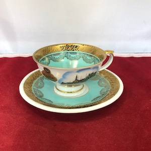 May include: A white teacup and saucer with a gold trim and a blue and white image of a cityscape. The teacup has a handle and the saucer has a scalloped edge.