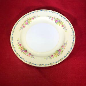 May include: A vintage floral design dinner plate with a cream colored background and a blue and gold trim. The plate features a delicate floral pattern with pink, yellow, and purple flowers.