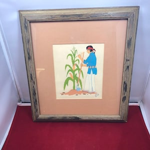 May include: A framed illustration of a person in traditional clothing tending to a corn plant. The person is wearing a blue shirt, white pants, and a red headband. The corn plant is green with yellow ears of corn. The illustration is set against a peach background.
