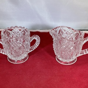 May include: A clear glass sugar bowl and creamer set with a cut glass design. The set has two handles and a round base.
